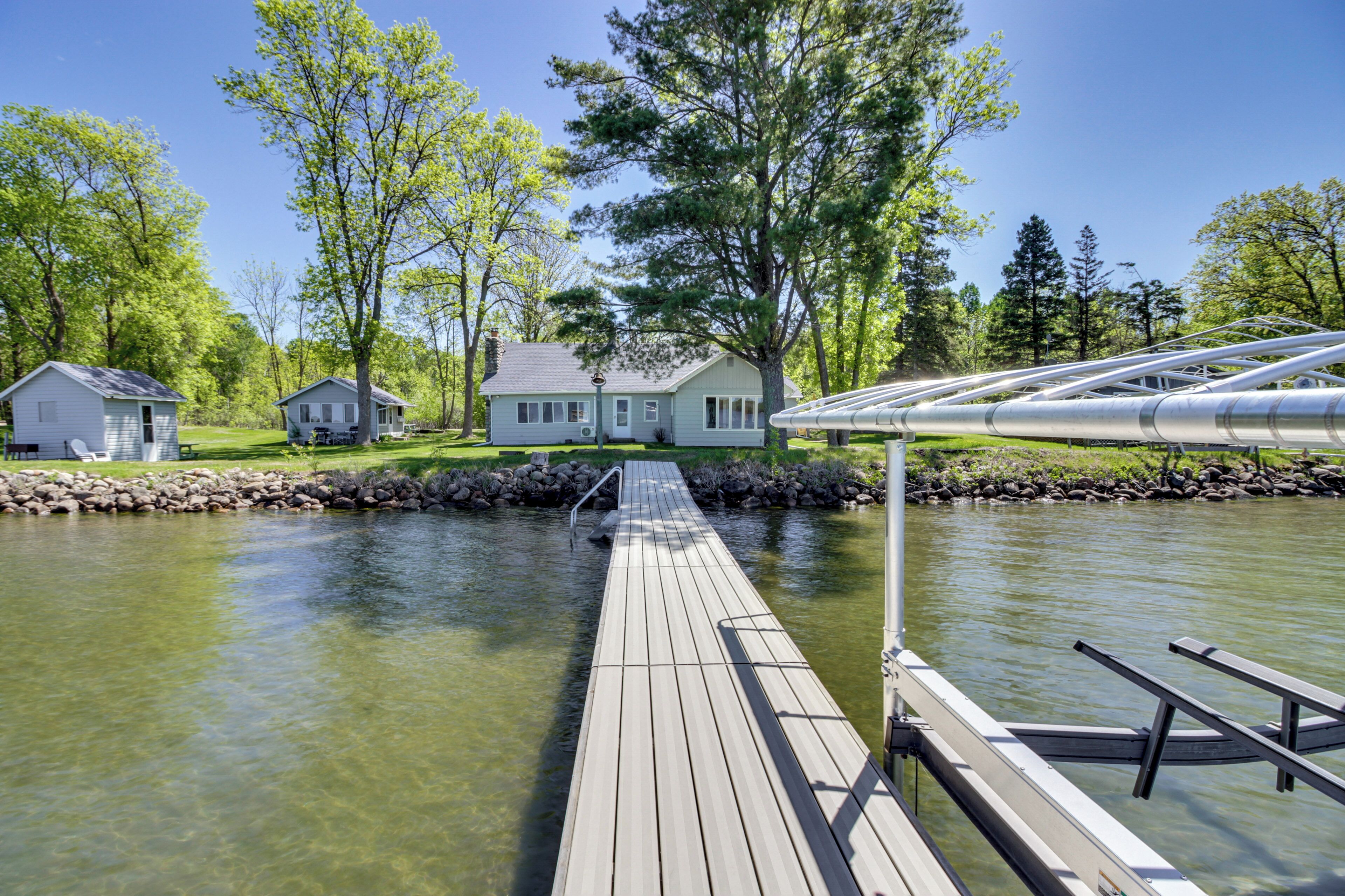 A wooden dock leading to a house by a calm river, surrounded by trees and a stone wall.