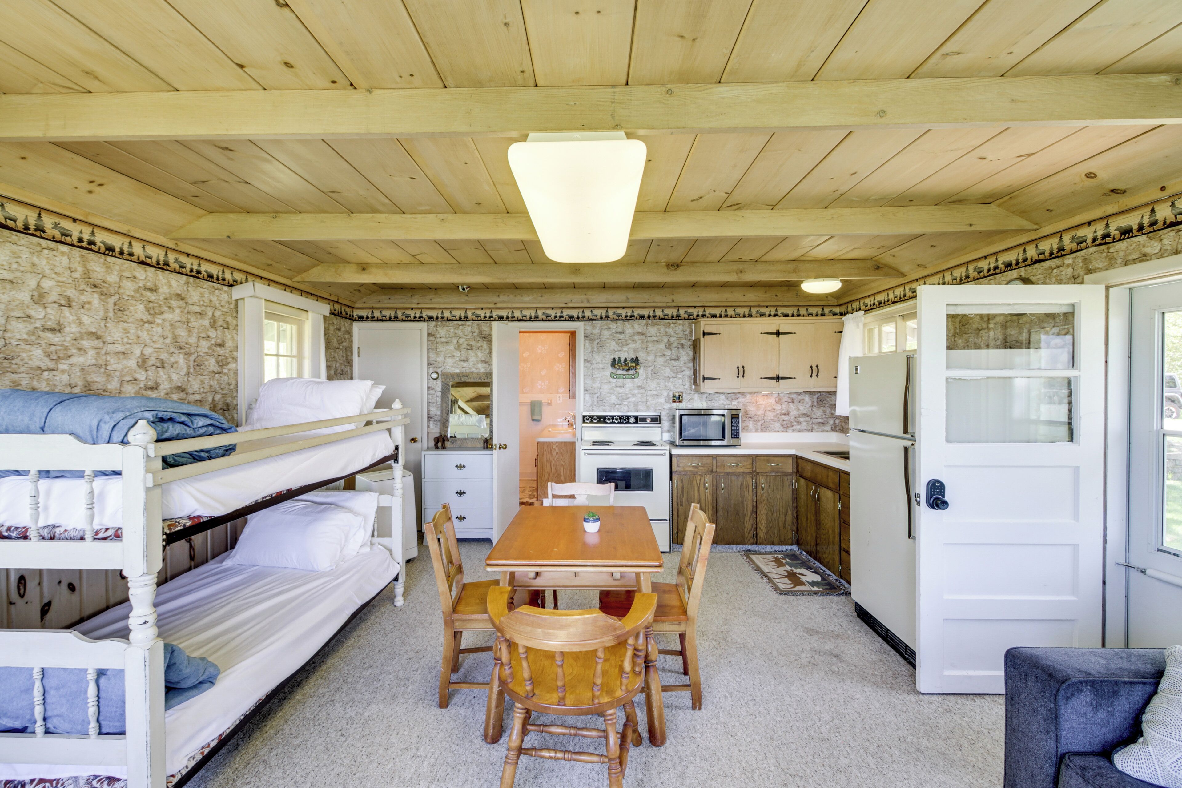 A compact room with bunk beds, a wooden dining table, chairs, a kitchen area with appliances, and a door leading to another room.