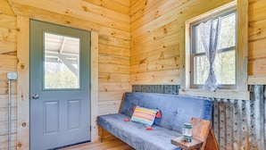 A wooden cabin interior with a gray door, a blue sofa with a colorful cushion, a small wooden table with a lantern, and a window with patterned curtains. - 'fins & Flowers' Off-grid Cabin w/ Trout Fishing! (Lewiston)
