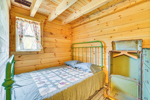 A wooden cabin room with a bed, a window with floral curtains, and a green metal bed frame. - 'fins & Flowers' Off-grid Cabin w/ Trout Fishing! (Lewiston)
