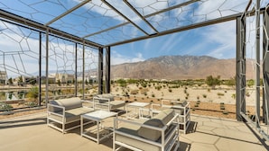 A modern outdoor lounge area with white furniture and a glass enclosure, offering a view of a desert landscape and mountains.
