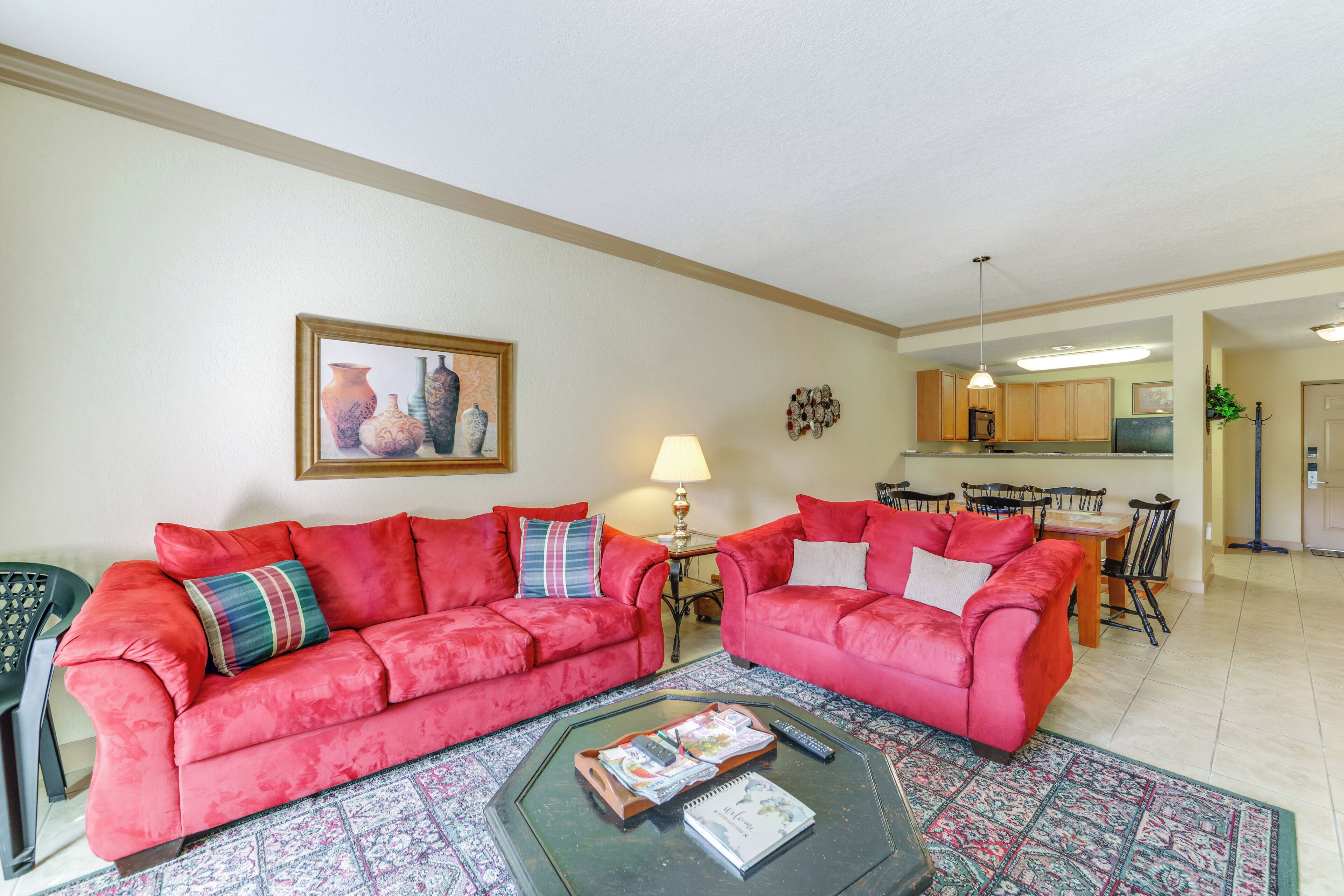 A living room with two red sofas, a coffee table with magazines, a lamp, and a kitchen in the background.