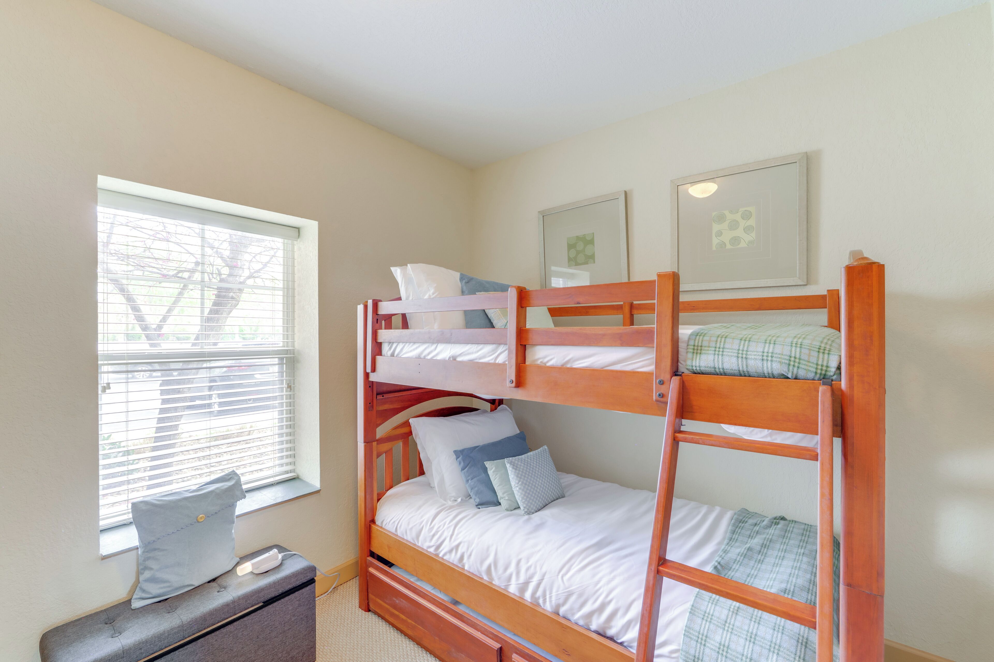 A bunk bed with two levels, a window with blinds, a bedside table, and two framed pictures on the wall.