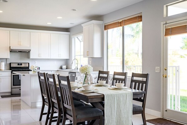 A modern kitchen with white cabinets, a dining table set for six, and a large window with blinds.