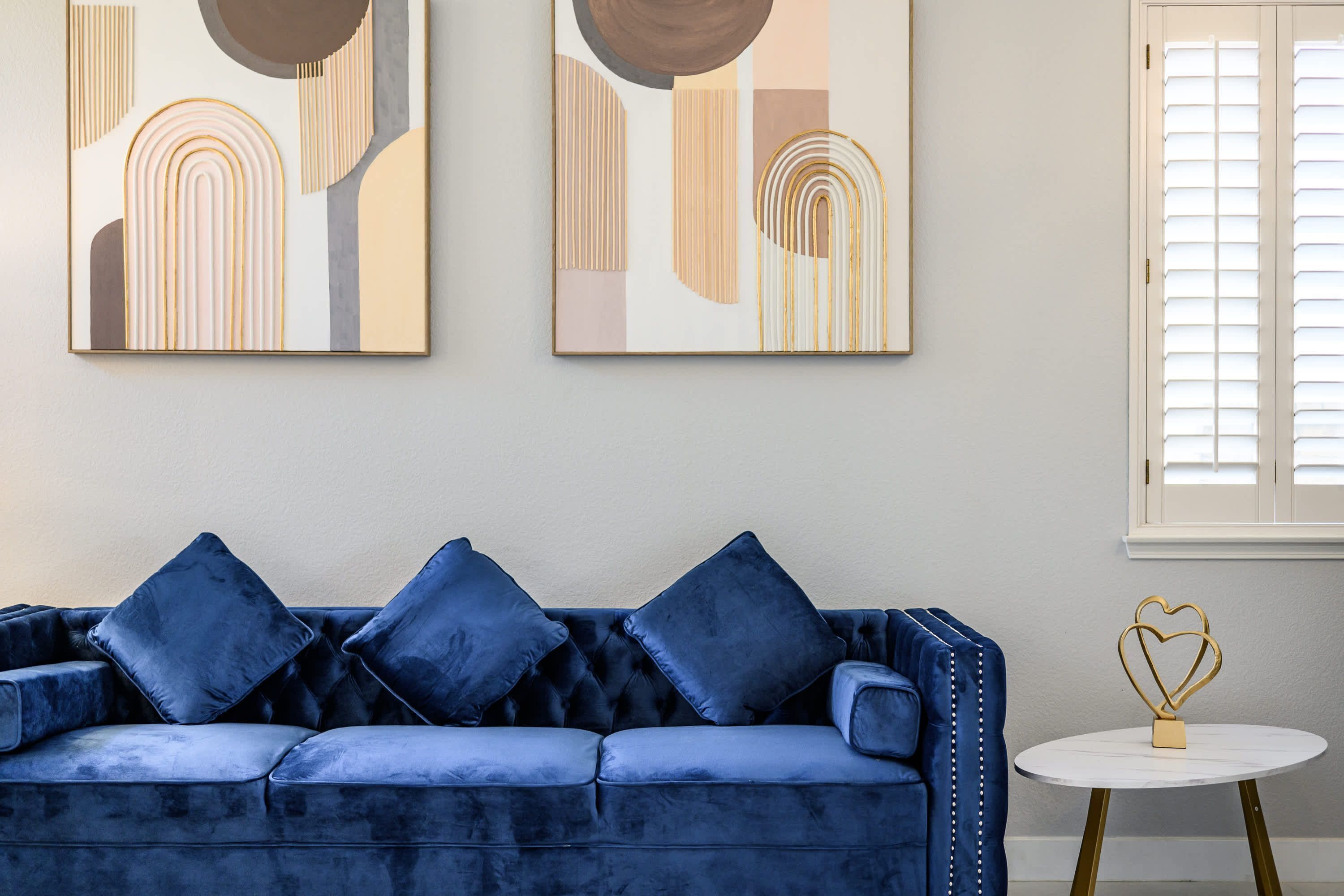 Un sofá de terciopelo azul con tres cojines, una mesa redonda dorada con un centro de mesa con forma de corazón y dos cuadros abstractos en la pared.