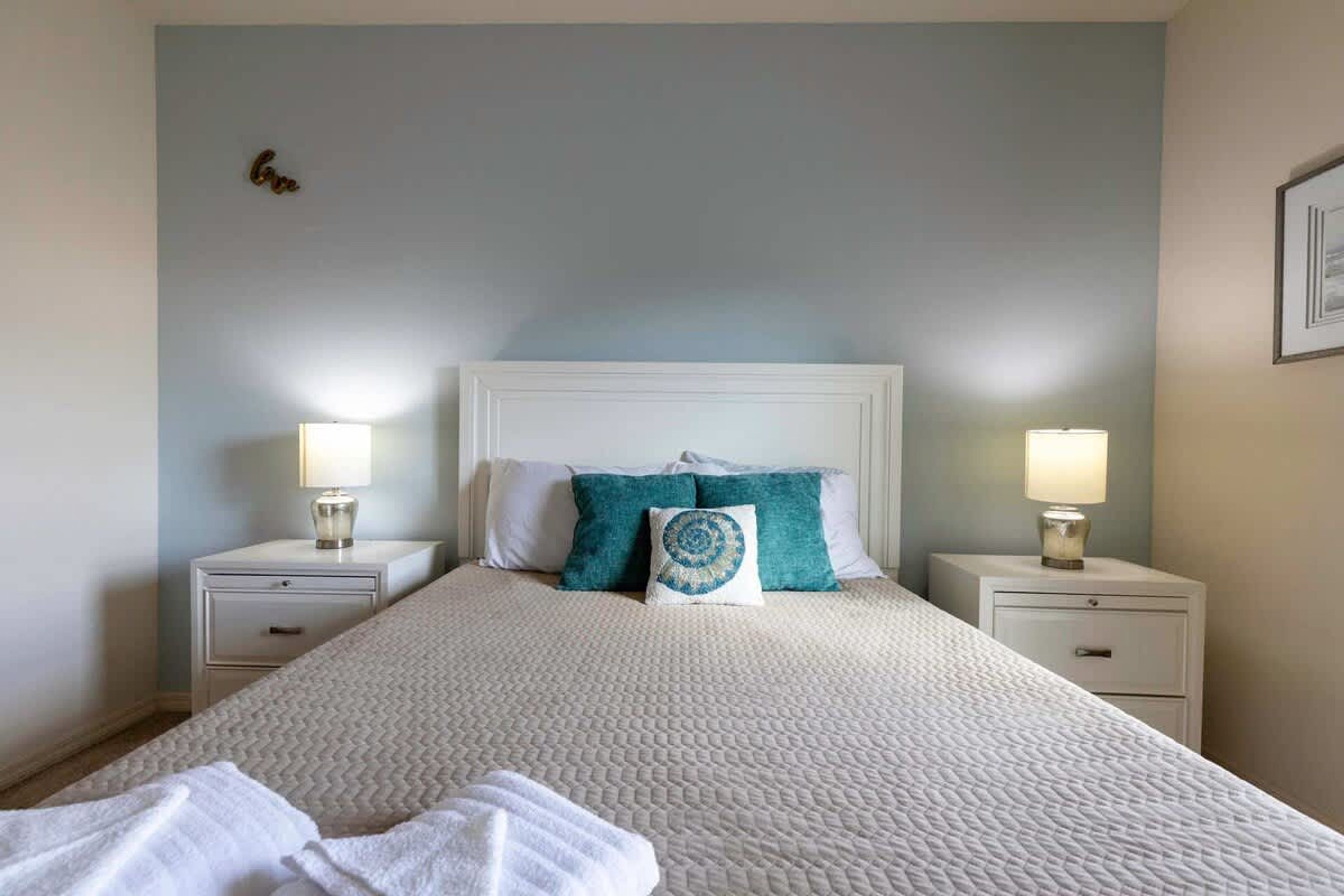 Een net opgemaakt bed met een beddegoed met patroon, twee nachtkastjes met lampen, en een muur met een klein decoratief object.