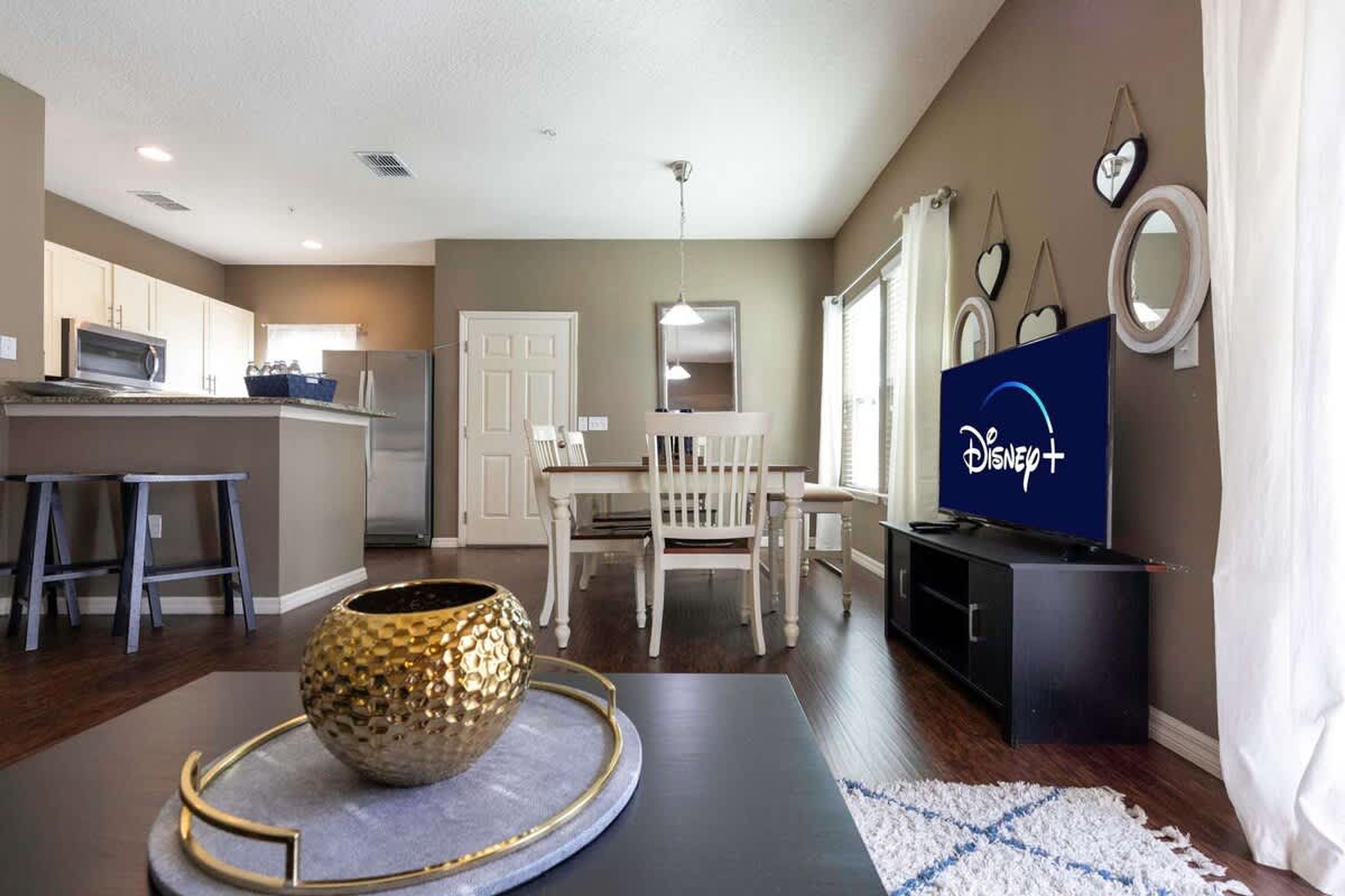 A modern living room with a dining area, a kitchen, and a TV displaying the Disney+ logo.