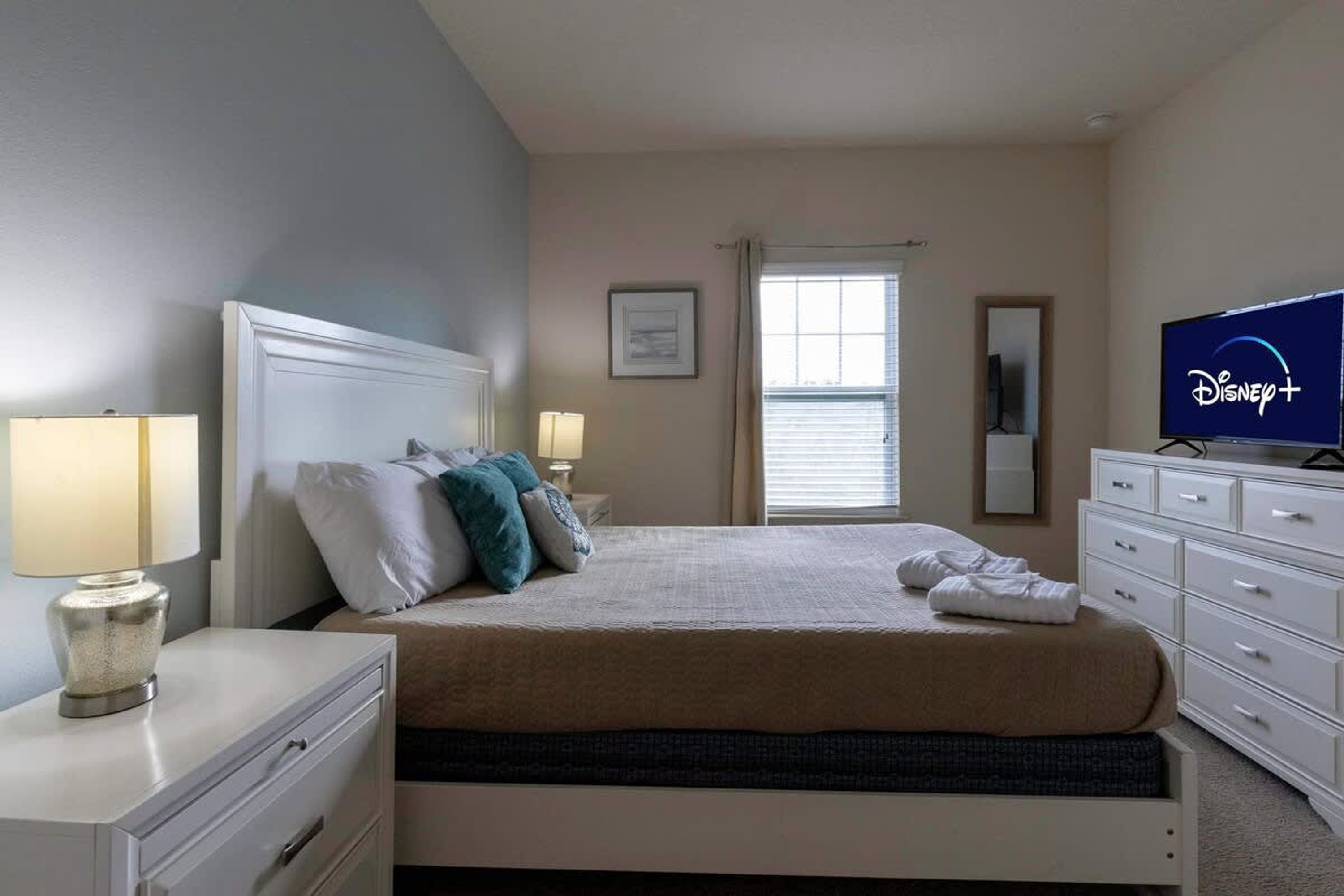 A hotel room with a bed, a TV displaying Disney+, a dresser, and a window with blinds.