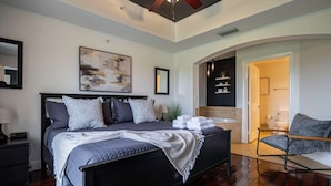 Un dormitorio con cama, ventilador de techo, un cuadro y baño visible a través de una puerta abierta.