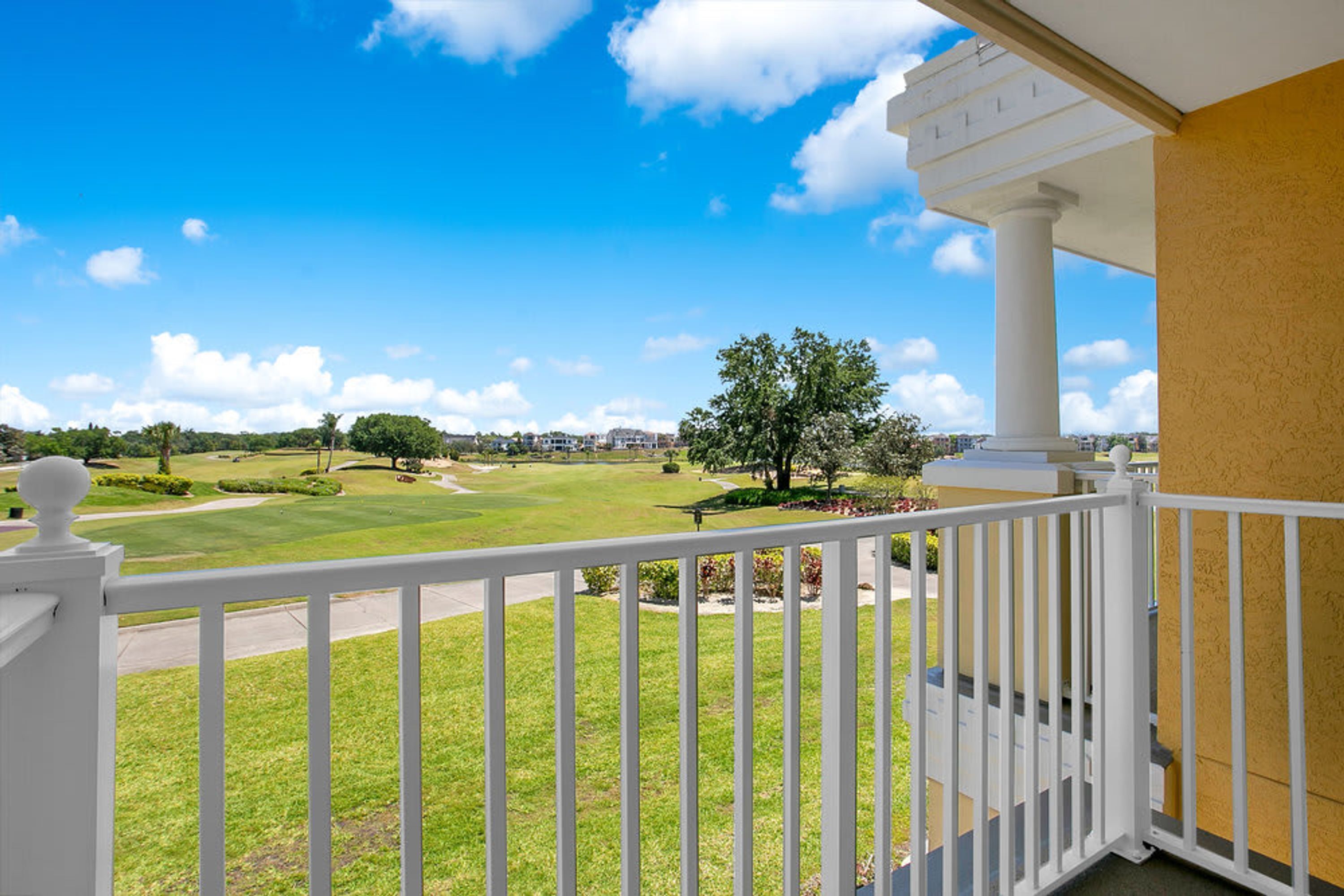 A balcony with a view of a golf course, white railings, and a clear blue sky.