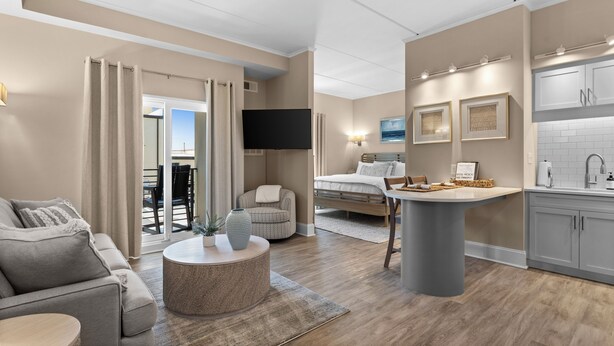 A modern living room with a sofa, coffee table, and a TV. There is a kitchen area with a sink and cabinets.