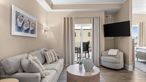 A living room with a sofa, armchair, and round coffee table, a flat-screen TV mounted on the wall, and a balcony with a view.