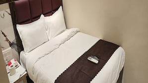 Un lit bien fait, recouvert d’une couverture blanche et marron, avec une télécommande posée dessus et une petite table de chevet sur laquelle se trouvent une boîte de mouchoirs et une bouteille.
