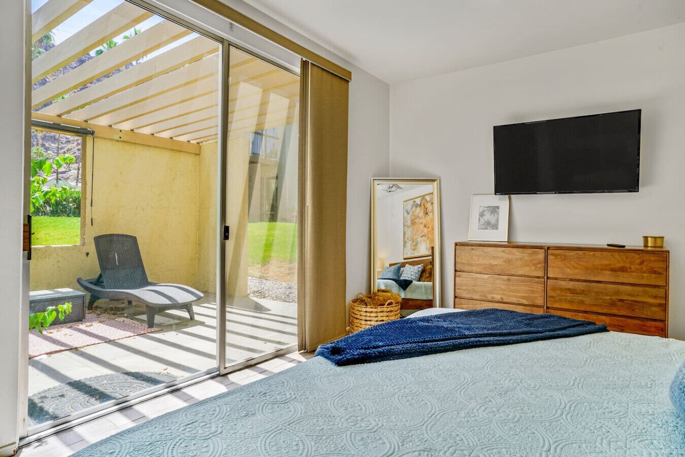 Une chambre avec un lit, une commode, un téléviseur et une porte-fenêtre coulissante donnant sur une terrasse extérieure avec une chaise et un panier.