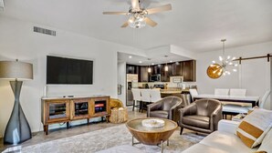 Un salon moderne avec un canapé, des fauteuils, une table basse, un téléviseur et, au fond, un coin cuisine.