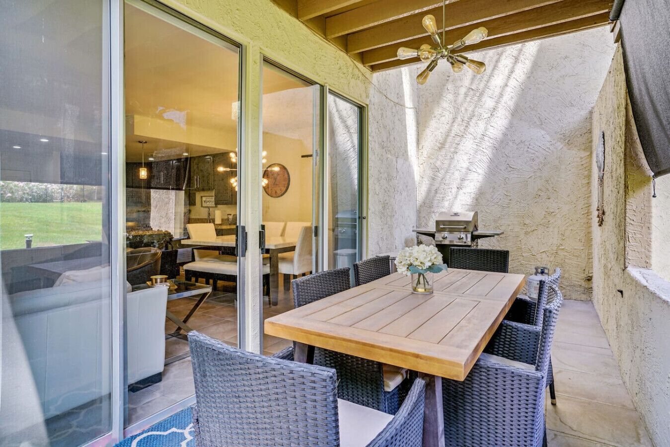 Un patio con mesa de madera, sillas de mimbre, una parrilla y vista al jardín trasero.