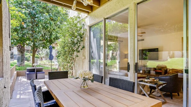 Un espace patio avec une table et des chaises en bois, un vase de fleurs et une porte vitrée donnant sur un jardin.