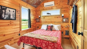 A wooden cabin room with a bed, a window with a view, and a mounted air conditioner. - Cozy Cottage w/ Firepit, Pickleball & Porch (Wimberley)