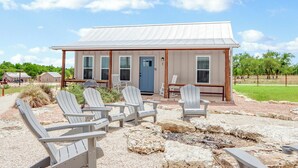 A rustic building with a metal roof, a blue door, and a patio area with Adirondack chairs. - Cozy Cottage w/ Firepit, Pickleball & Porch (Wimberley)