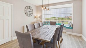 A dining area with a wooden table, grey upholstered chairs, a clock, and a view through large windows. - Host & Stay | Chirnells (Bamburgh)
