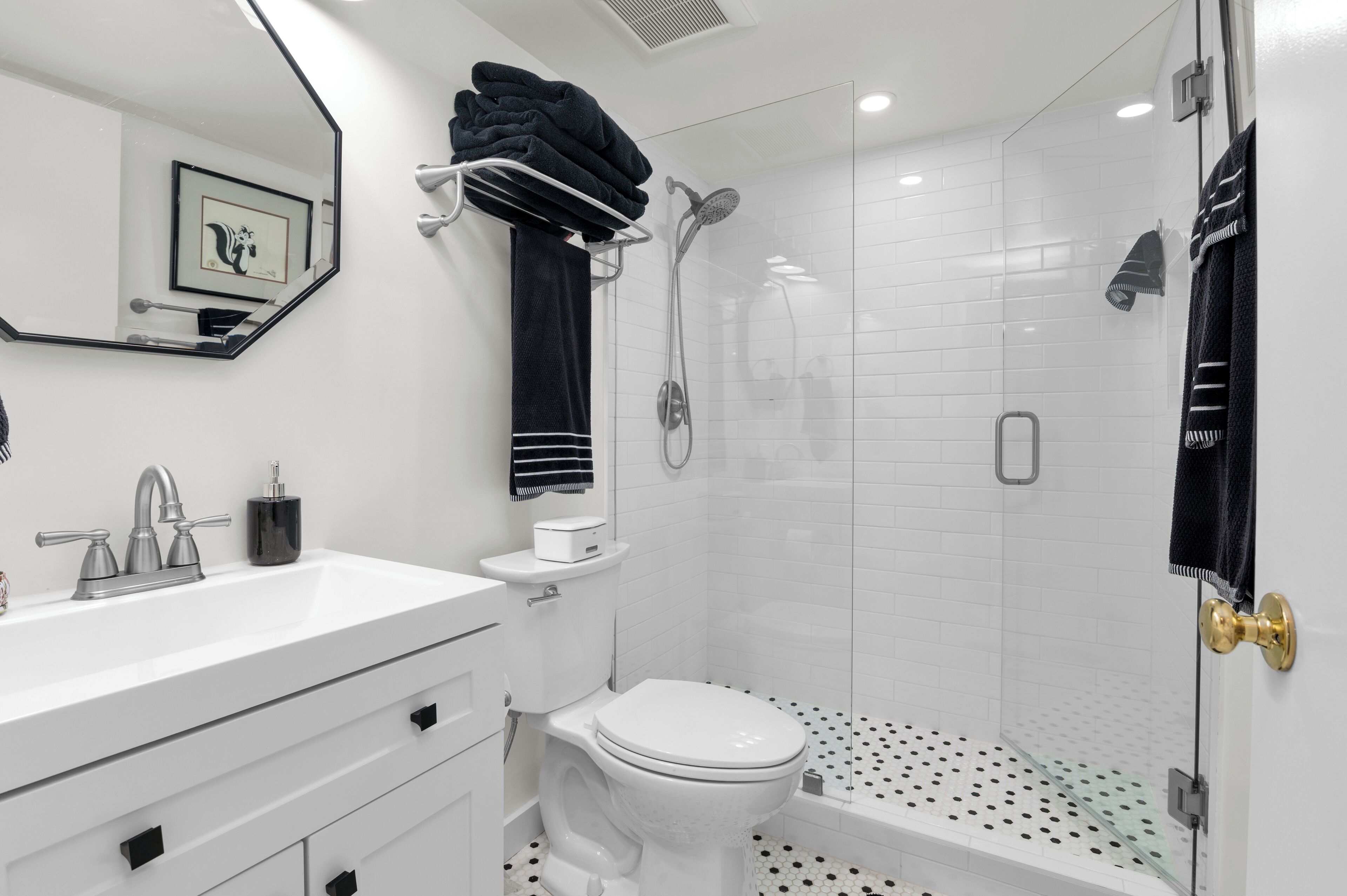 Een moderne badkamer met een witte wastafel, toilet en douche. Er is een handdoekhouder met opgevouwen handdoeken en een ingelijst schilderij aan de muur.