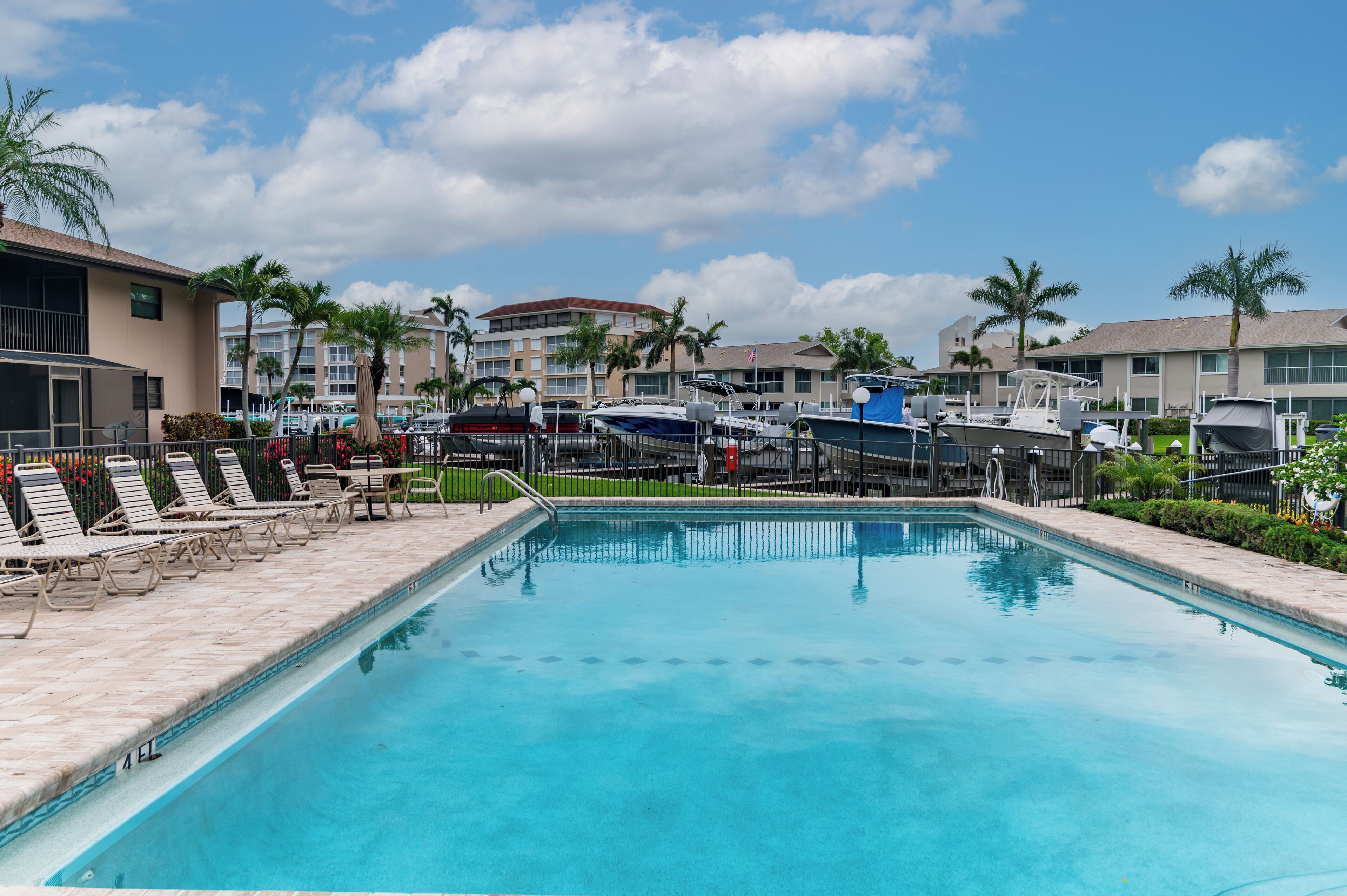 A swimming pool with lounge chairs, boats, and a hotel building in the background.