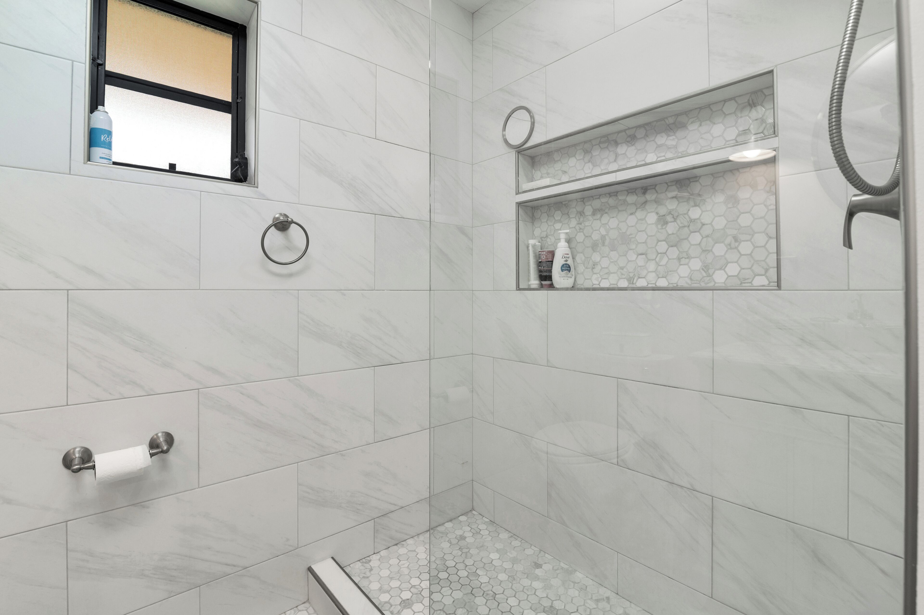A modern bathroom with white marble tiles, a glass shower enclosure, a towel rack, and a shelf with toiletries.