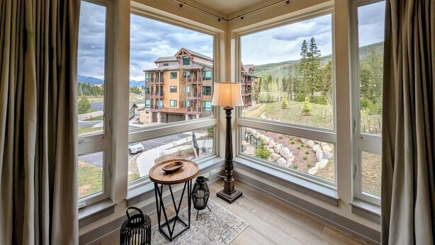 A room with large windows, a table, a lamp, and a view of a building and landscape. - Views & Trails | Updated Retreat Summer Activities (Keystone)