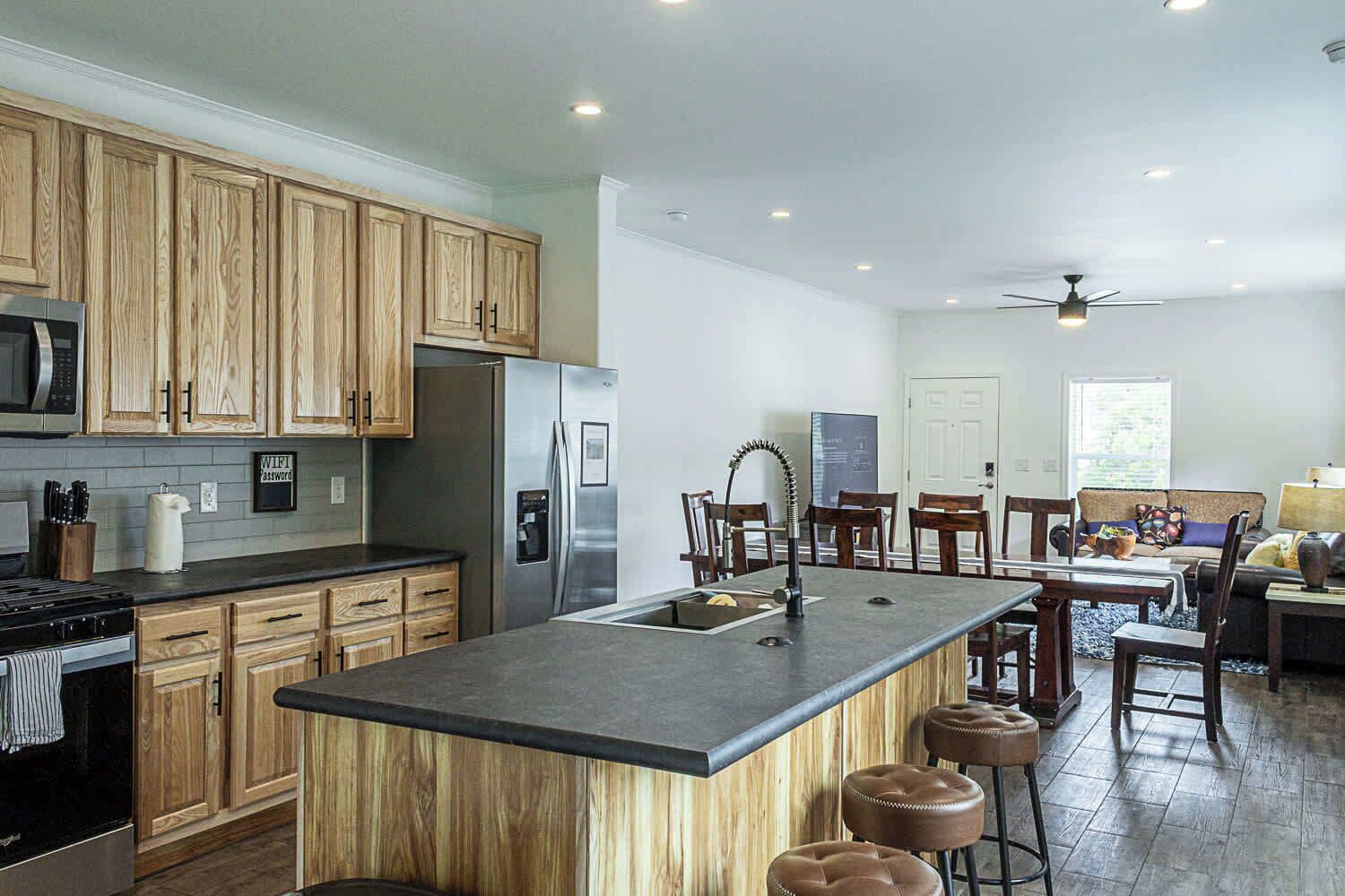 Una cocina moderna con armarios de madera, electrodomésticos de acero inoxidable, una isla central con taburetes y un comedor con ventilador de techo.