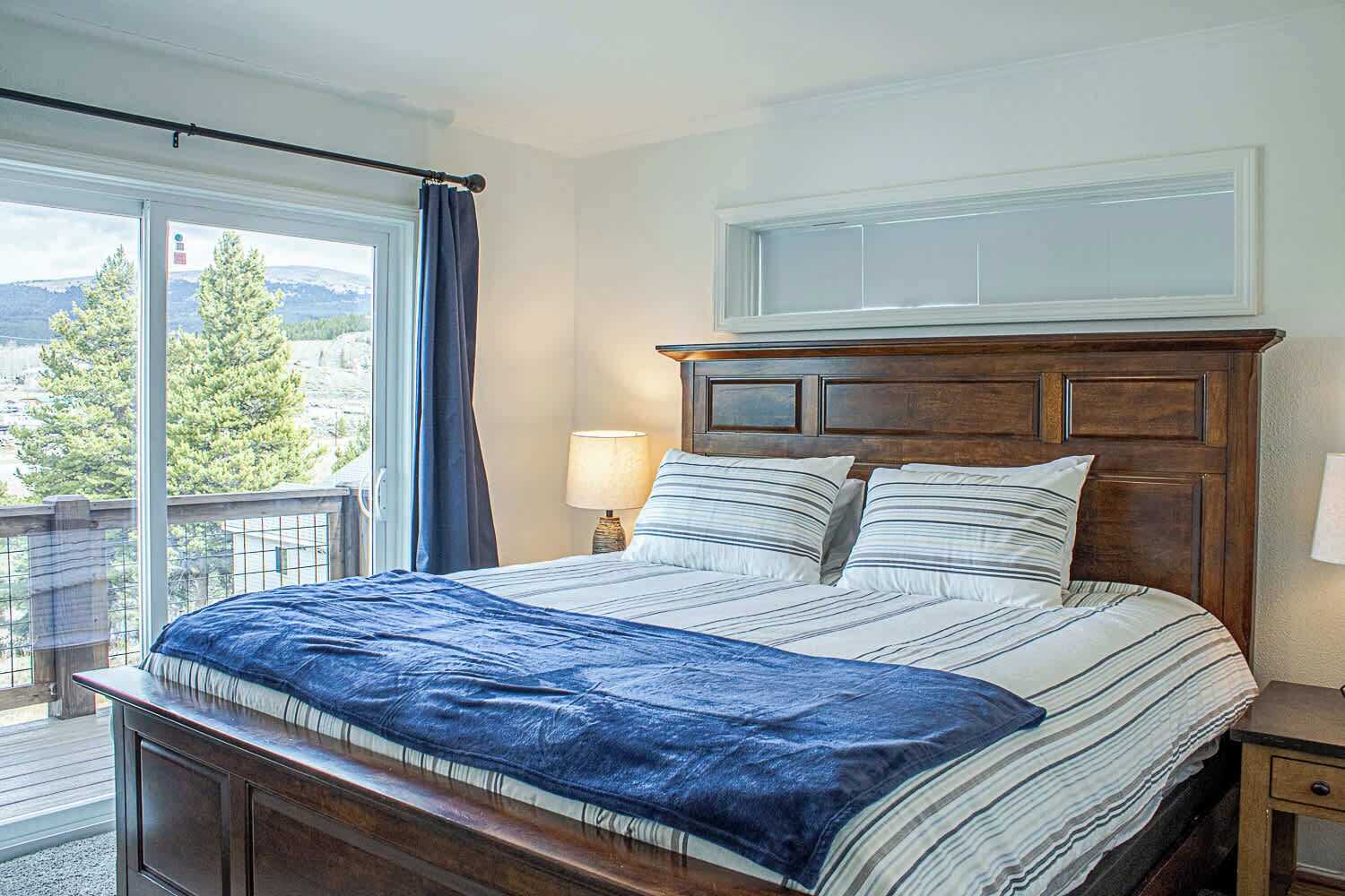 Ein Schlafzimmer mit einem großen Bett, einem hölzernen Kopfteil, einem Nachttisch, einer Lampe und Blick auf Bäume und Berge durch ein Fenster.