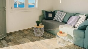 A living room with a sofa, a coffee table, and a view of the sea through a window. - Seafront Alos Gem - Loutra Seaview Escape (Loutra)