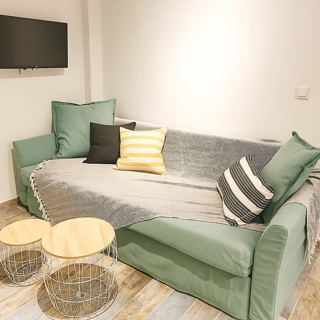 Un sofá verde con una manta estampada, dos mesas redondas de madera y un televisor de pantalla plana montado en la pared.
