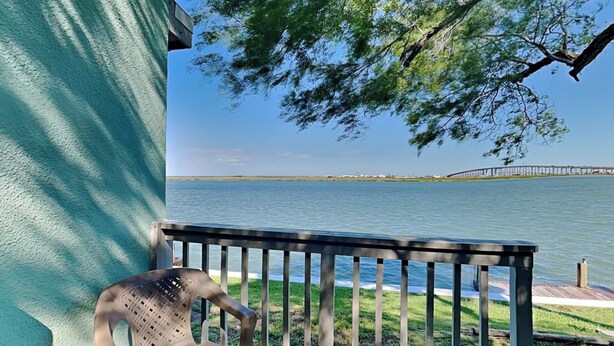 A wooden deck with a chair, overlooking a calm body of water and a distant pier. - Watercolor Skies | Cozy, Waterfront (Corpus Christi)