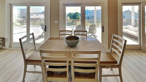 A dining area with a large table and six chairs, a bowl on the table, and a view of a deck and outdoor furniture through glass doors. - Papa's Paradise | Partial Beach Views (Galveston)