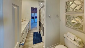A narrow hallway with a wooden floor, a white toilet, and a shower area with a checkered curtain. - Papa's Paradise | Partial Beach Views (Galveston)