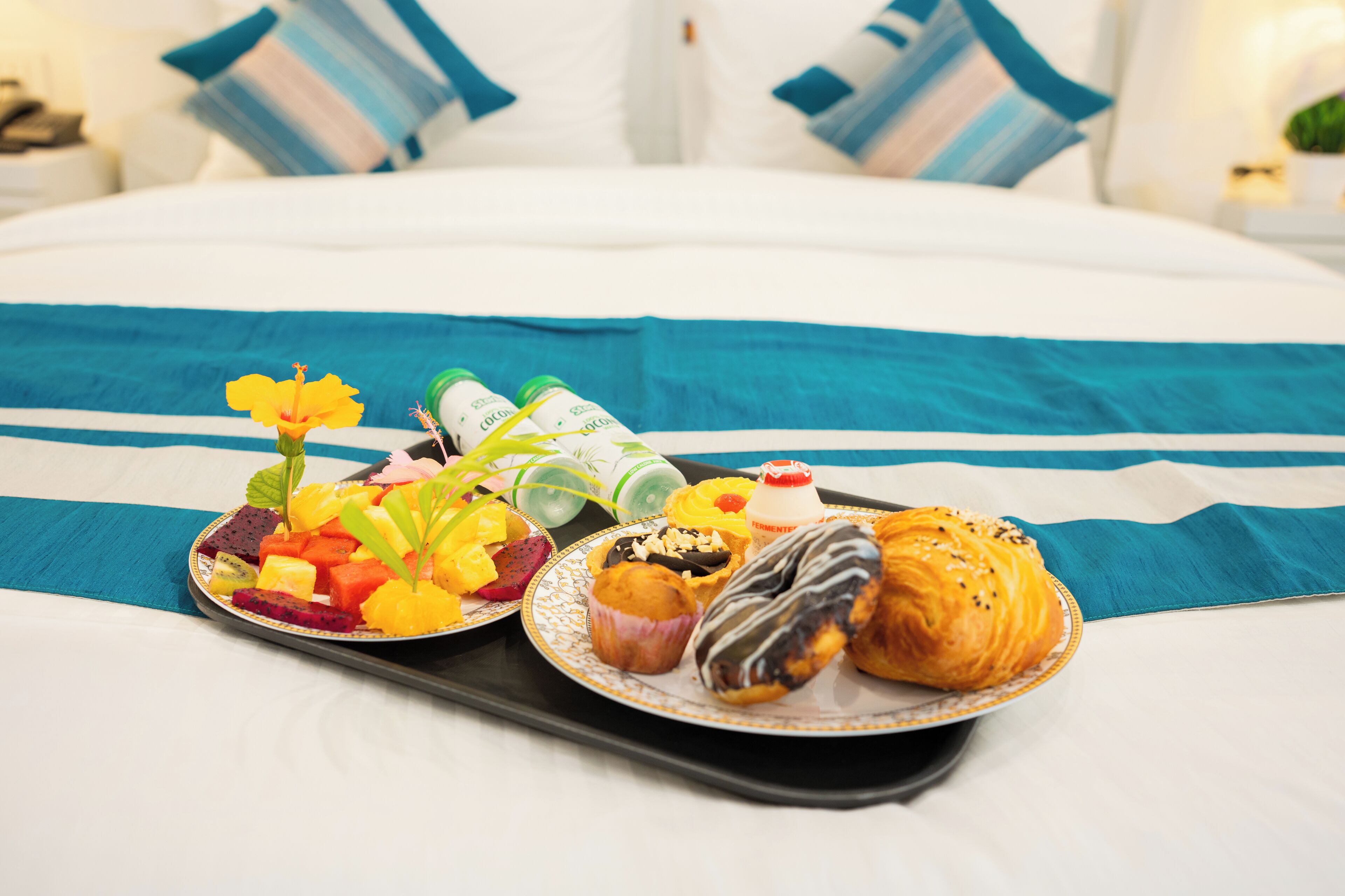 Una bandeja con alimentos para el desayuno, como frutas, muffins y un croissant, sobre una cama con colcha azul y blanca a rayas.
