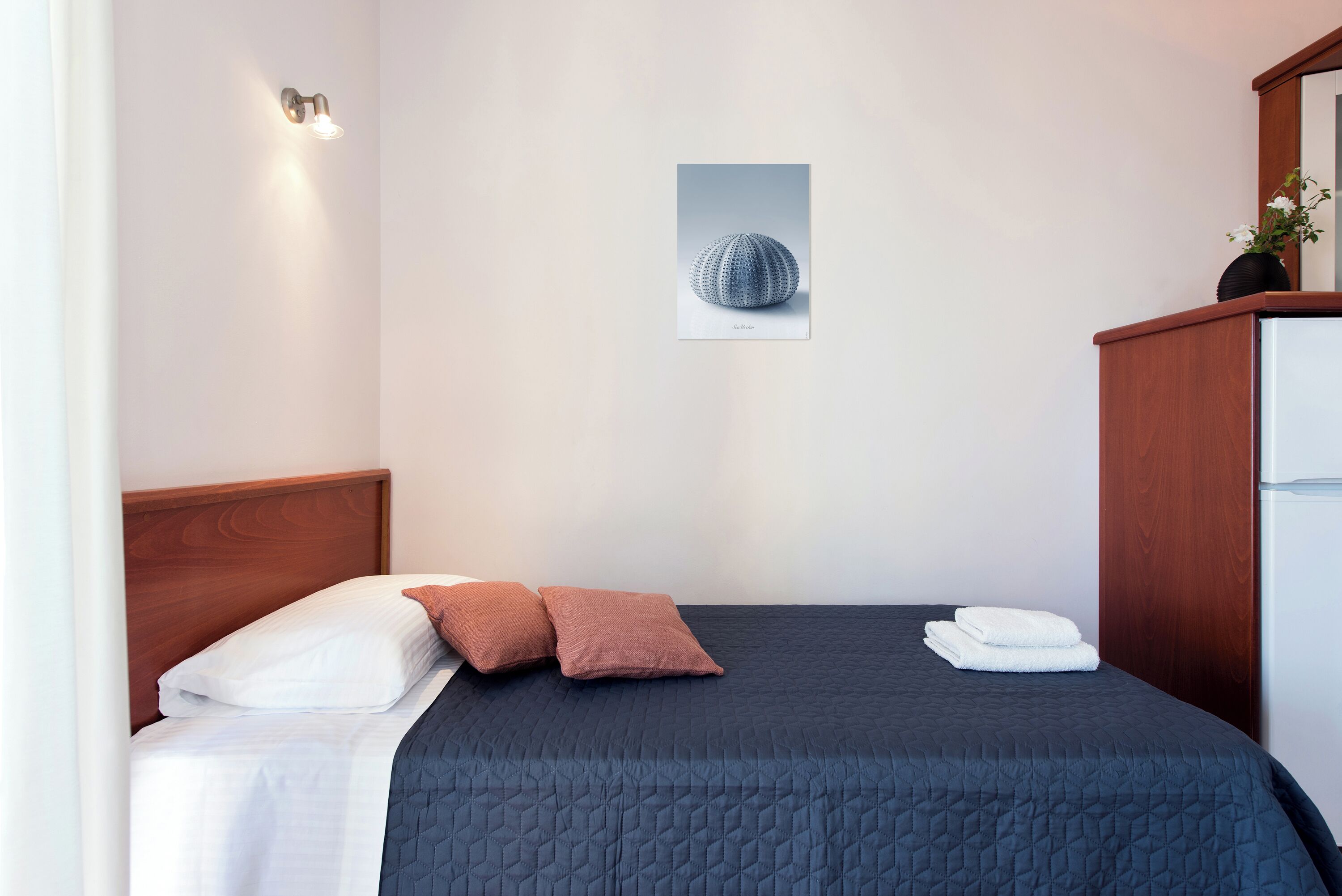 Una cama bien tendida con un edredón oscuro, dos almohadas y una manta blanca. Un cuadro enmarcado cuelga de la pared sobre la cama.