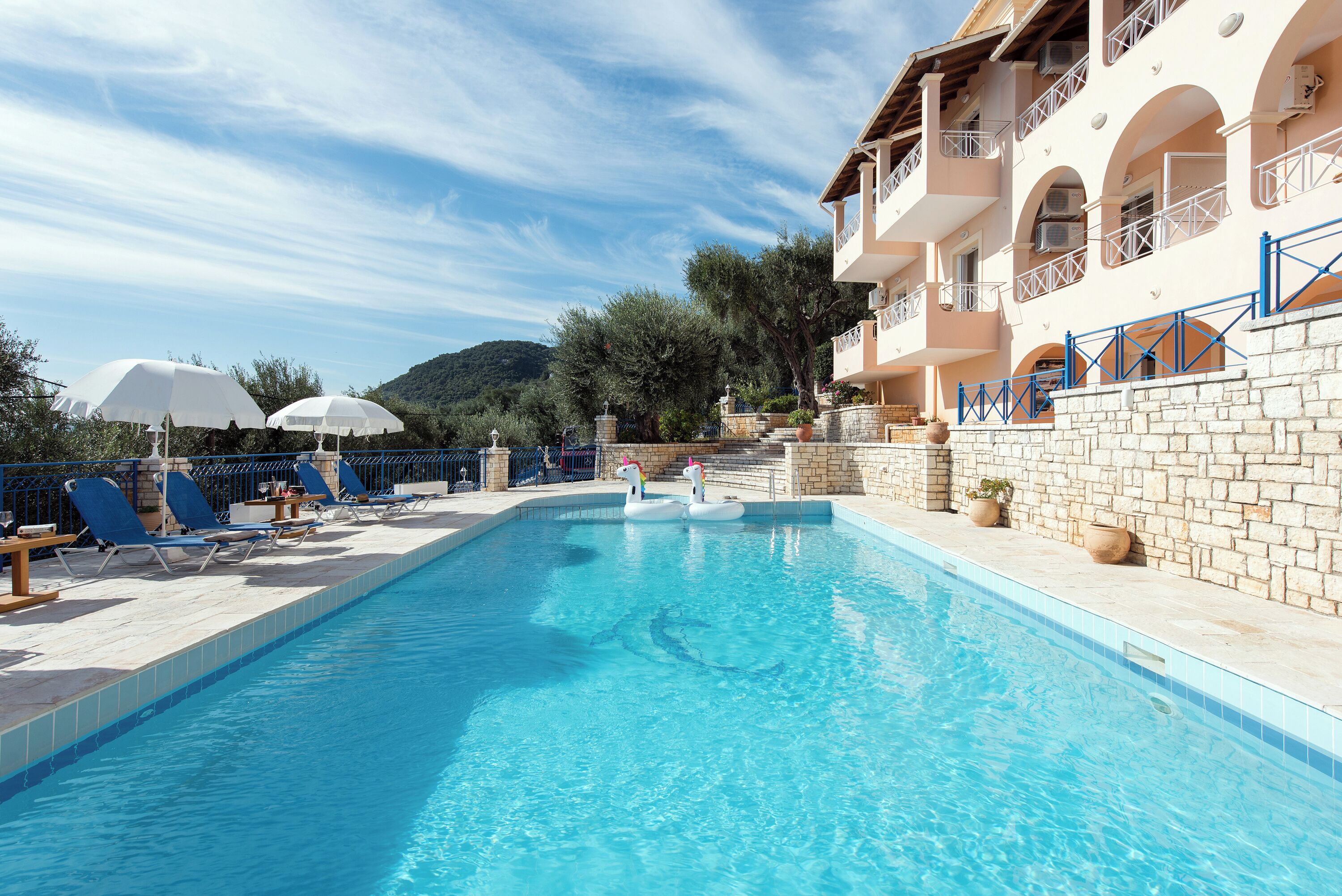 Une piscine entourée de chaises longues et de parasols, le tout bordé de murs en pierre et adossé à un bâtiment doté de balcons.