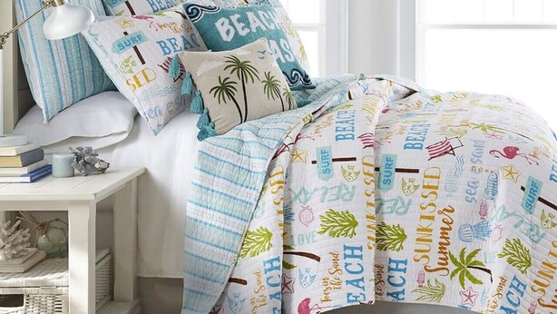 Un dormitorio con una cama cubierta por un edredón con motivos playeros, una mesita de noche con libros y una lámpara, y una ventana que ofrece vistas al cielo.