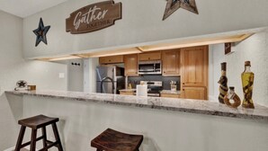 Una cocina con armarios de madera, un refrigerador de acero inoxidable y una encimera de mármol. Hay dos taburetes y letreros decorativos con forma de estrella en la pared.