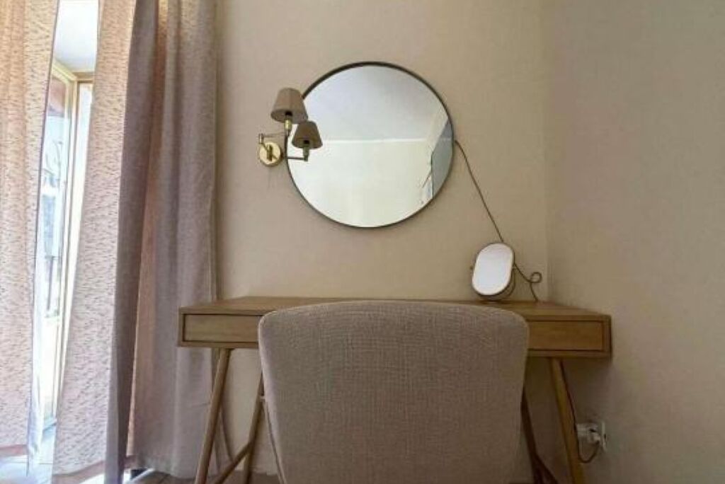 Una habitación con un espejo redondo en la pared, un escritorio con una lámpara y una silla.