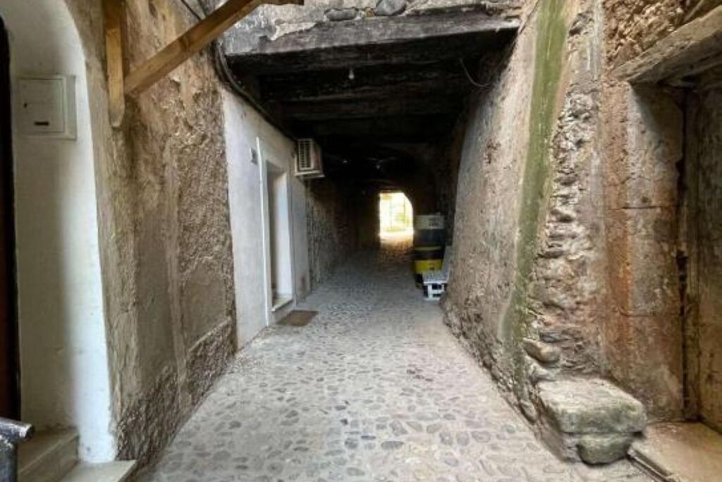 A narrow stone corridor with a rough, uneven floor, leading to a doorway at the end.