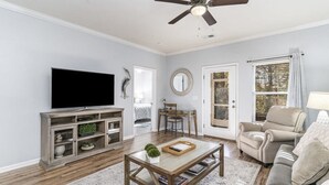 Un salon équipé d’un téléviseur à écran plat, d’un ventilateur de plafond, d’une table basse, d’un canapé et d’une fenêtre avec des rideaux.