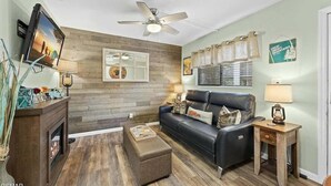 A living room with a fireplace, a black leather sofa, a wooden coffee table, and a ceiling fan. - Slopeside Serenity 111–Cabin Vibe–Sleeps 4 (Gatlinburg)