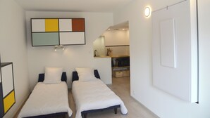 Una habitación de hotel moderna con dos camas individuales, una kitchenette pequeña y un baño con espejo.