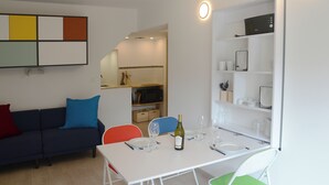Una sala moderna con una mesa de comedor blanca para cuatro personas, un sofá azul con cojines de colores y una zona de cocina con armarios blancos y un microondas.