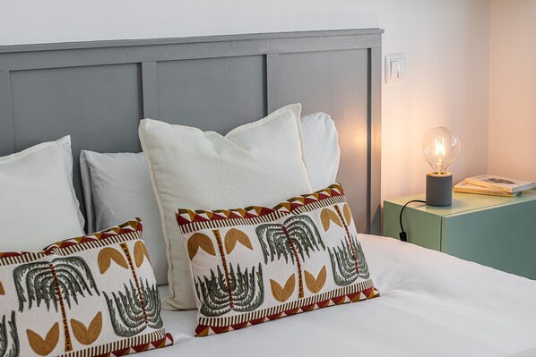 A neatly made bed with white bedding and decorative pillows, a gray headboard, and a bedside table with a lamp and a book.