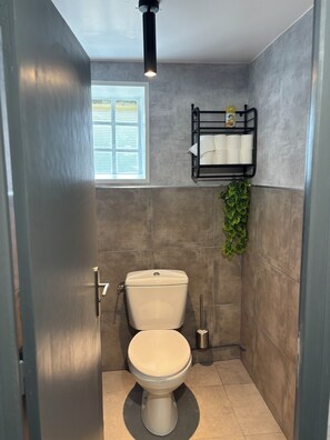 Une petite salle de bain comprenant des toilettes, une étagère avec du papier toilette, une fenêtre avec des stores et une petite plante.