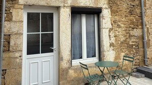 Un bâtiment à deux étages avec une façade en pierre, une porte blanche, une fenêtre avec un rideau et un petit espace extérieur aménagé avec une table et des chaises.