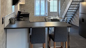 Une cuisine moderne dotée d’un îlot central, de deux tabourets de bar, d’une table à manger avec des chaises, d’un four encastré, d’un réfrigérateur en acier inoxydable et d’un escalier agrémenté d’un mur en pierre.